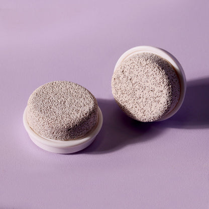 Two-pack of Cora Pumice Stone Replacement Brush Heads for facial cleansing and foot smoothing on a purple background.