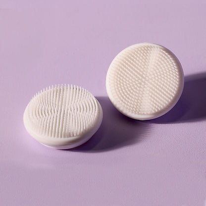 Two white silicone replacement facial cleansing brush heads for Cora devices on a purple background.